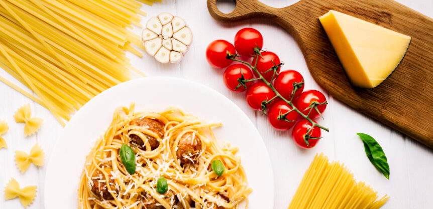 Piatto di spaghetti con polpette e parmigiano accompagnato da ingredienti crudi come pomodori ciliegino, aglio, pasta cruda, olive e una fetta di formaggio su un tagliere di legno.
