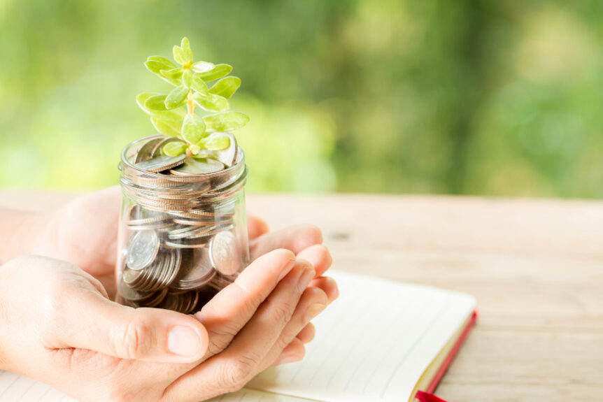 Mani che tengono un barattolo di vetro pieno di monete con una pianta che cresce su di esse, simbolo di investimento e crescita finanziaria.